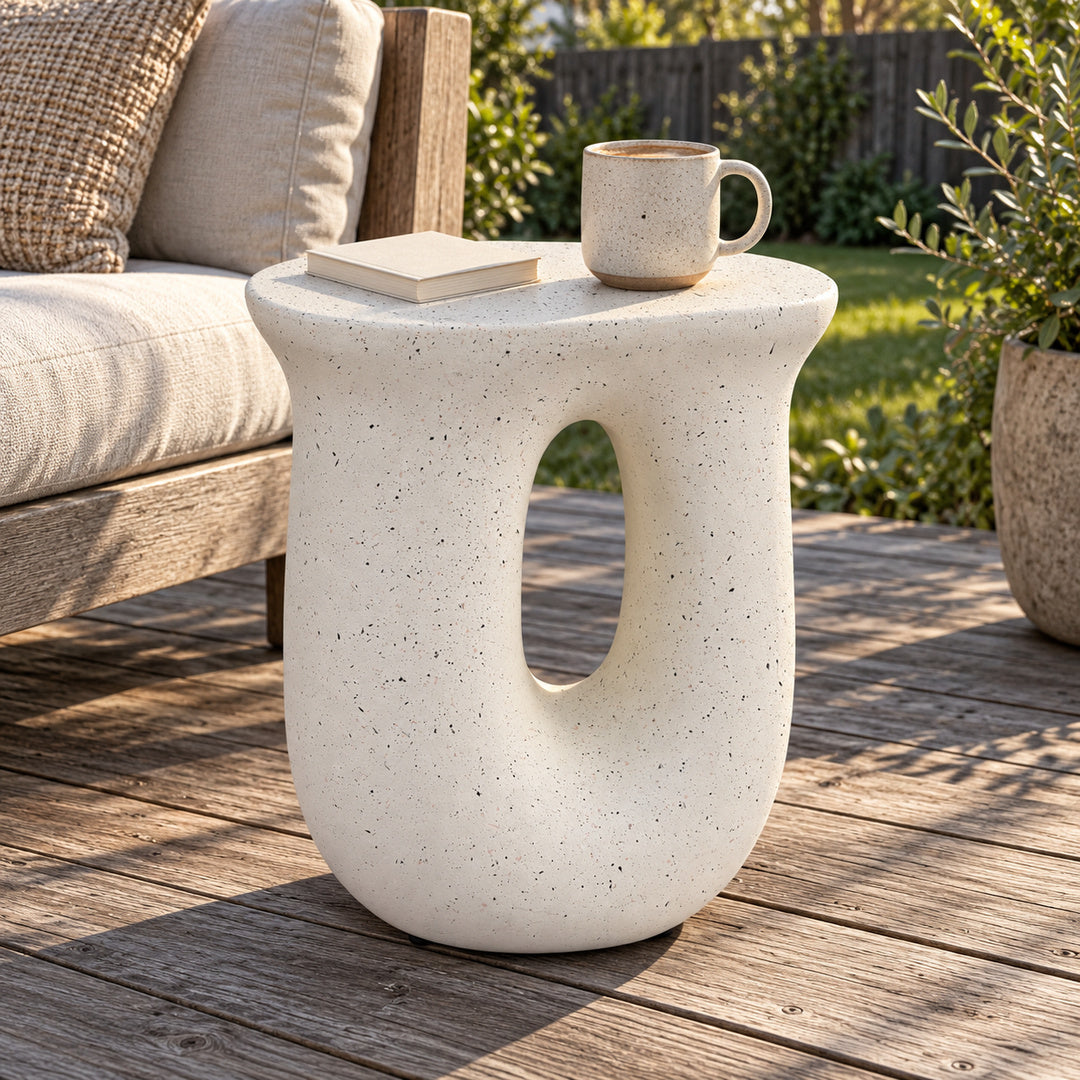 A modern U-shaped side table in a speckled off-white concrete finish, styled on a wooden deck next to a sofa with a mug and book on top.