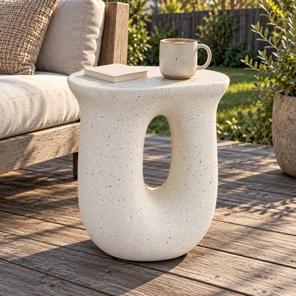 A modern U-shaped side table in a speckled off-white concrete finish, styled on a wooden deck next to a sofa with a mug and book on top.