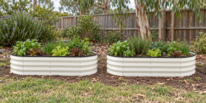Two corrugated metal raised garden beds filled with green herbs and plants, set in a backyard on grass and soil with a wooden fence and trees behind them.